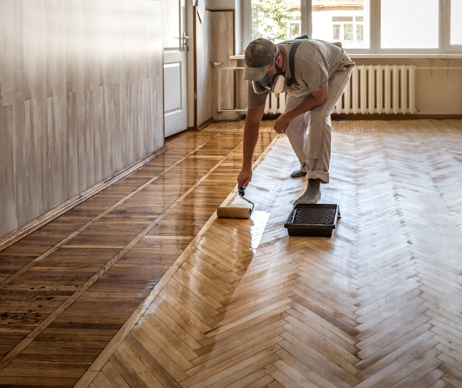 A person wearing a face mask applies varnish to a herringbone patterned wooden floor using a roller.